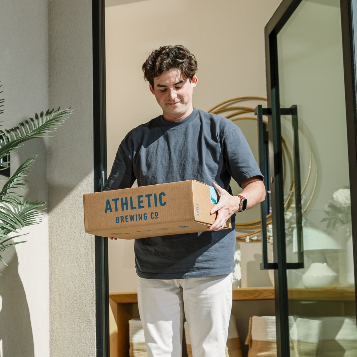 Man holding a box from Athletic Brewing Co. in a home setting