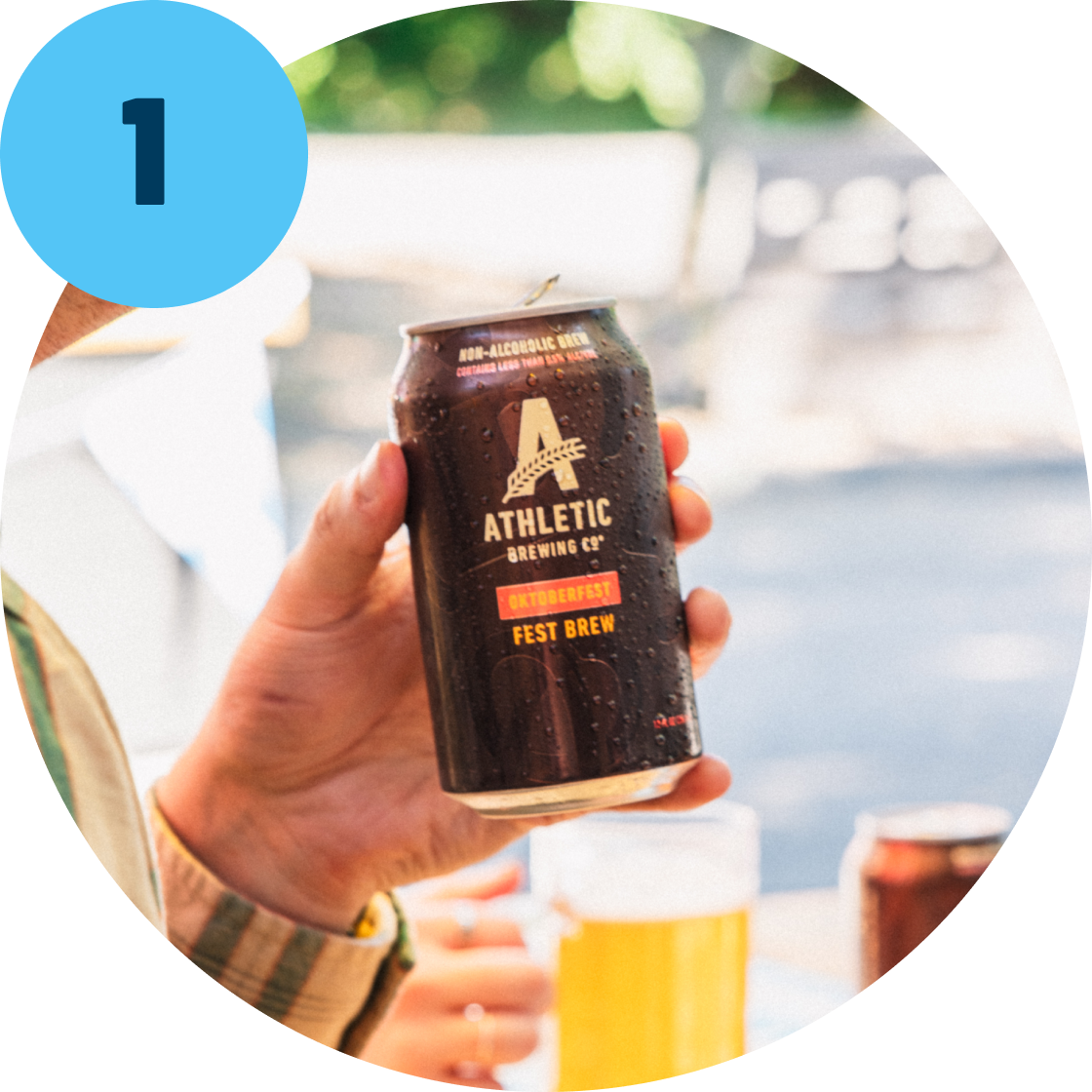Step 1 Hand holding a can of Athletic Brewing Co. non-alcoholic fest brew with a blurred outdoor background